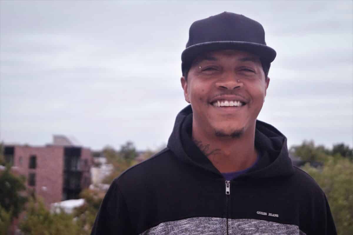 Smiling man wearing black Guess Jeans hoodie and cap, with urban buildings in background