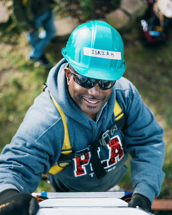 Worker named Isaiah in turquoise hard hat smiling at camera during construction work