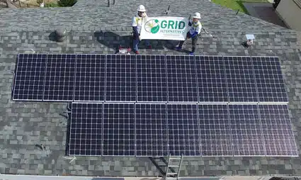 Two workers hold a GRID banner while standing on a residential roof with two solar panel arrays installed