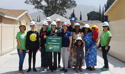 Group of volunteers in hard hats and colorful shirts posing with a solar panel installation sign on a residential roof