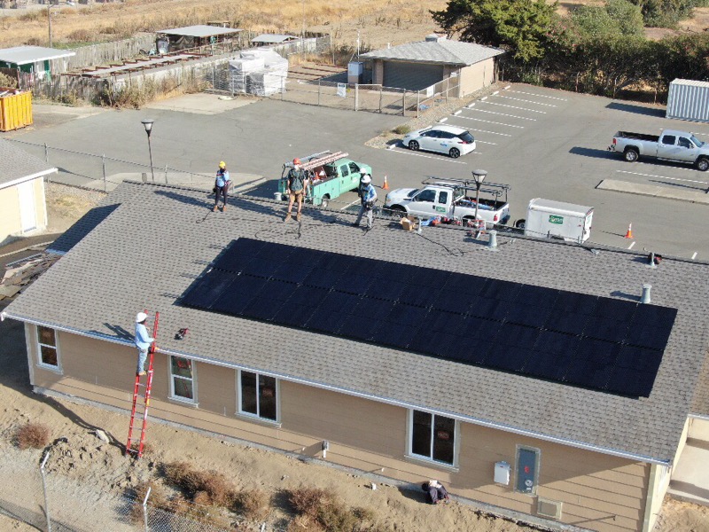 Solar panel installation on residential roof with work crew and vehicles in parking lot below
