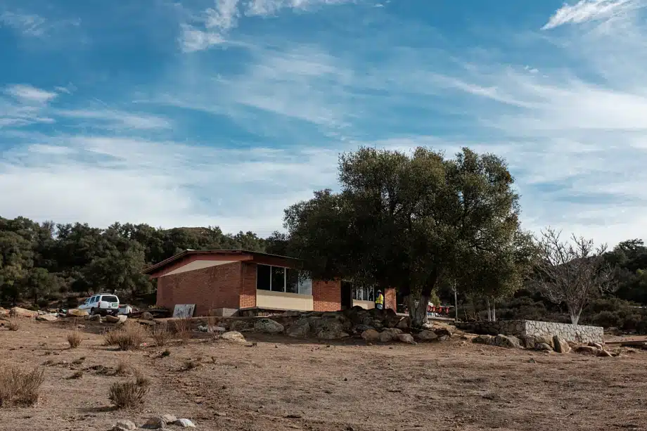 Brick house with red metal roof in arid landscape with scattered trees and boulders