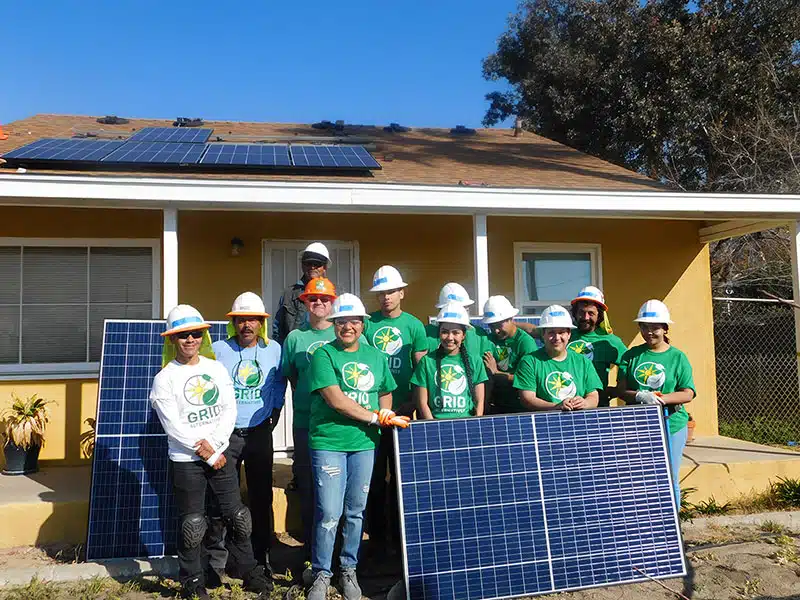 Group of GRID volunteers in green shirts and hard hats with solar panels at a yellow house installation
