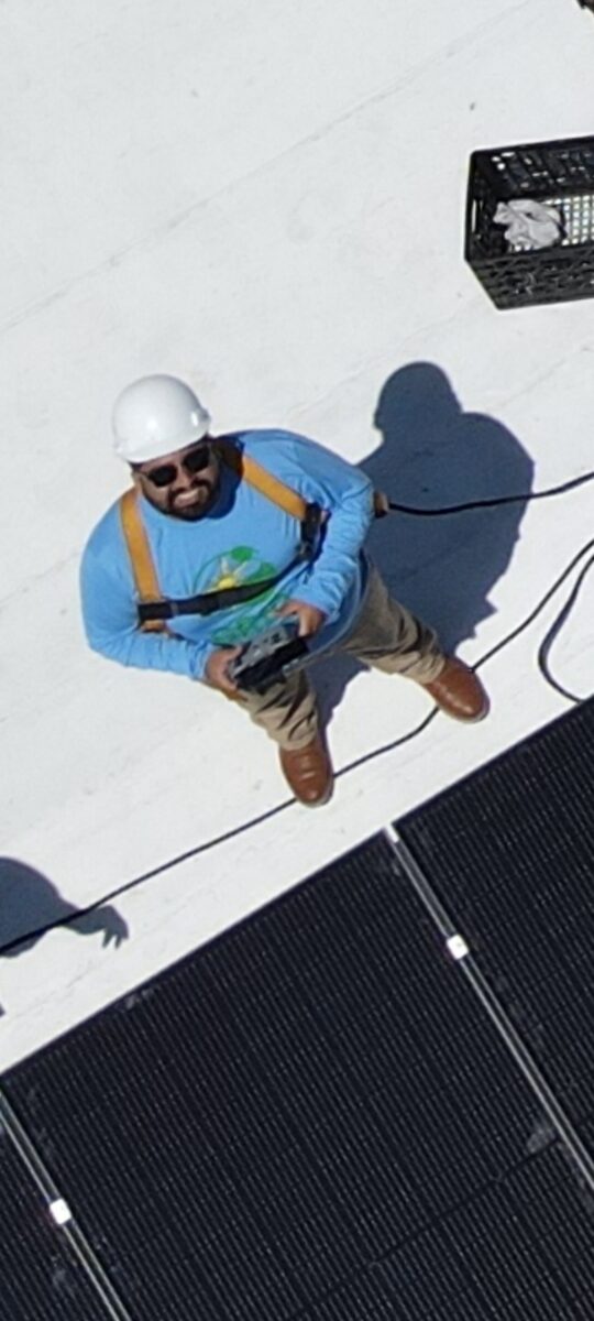 Jose Montes looking up and smiling on a rof next to a solar panel