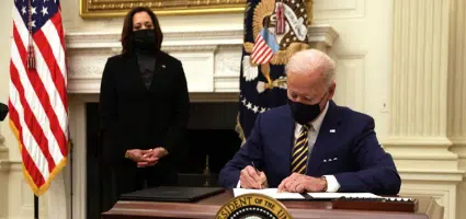 Man in suit signing document at Resolute desk with woman standing nearby; American flags visible