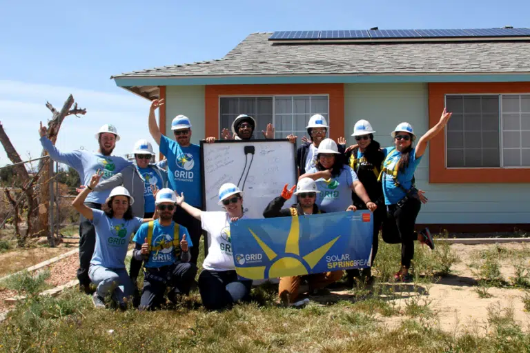 Team in hard hats celebrating in front of solar-powered house with "Solar Springbreak" banner