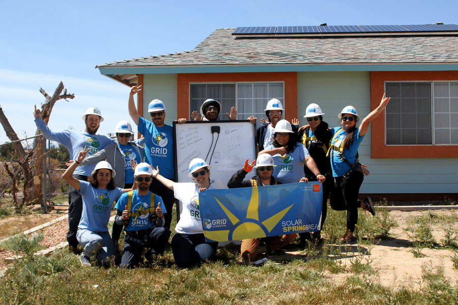 Team in hard hats celebrating in front of solar-powered house with "Solar Springbreak" banner