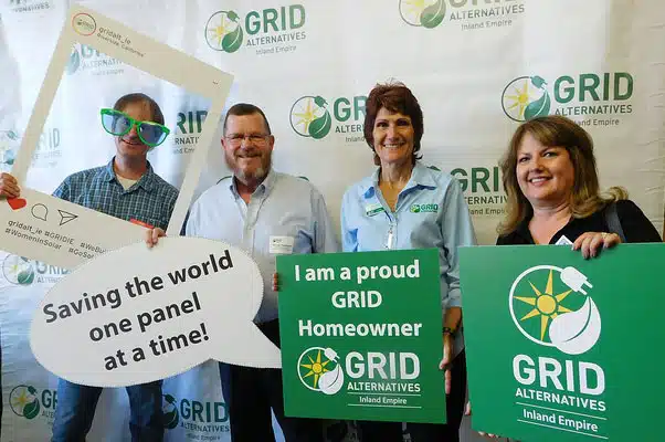 Four people holding GRID Alternatives signs at a solar event, with tagline "Saving the world one panel at a time