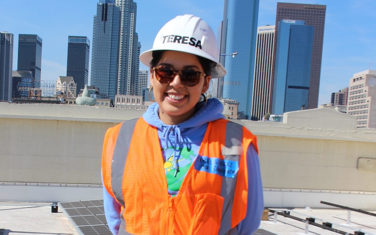 Woman in hard hat and safety vest smiling on rooftop with city skyline behind her