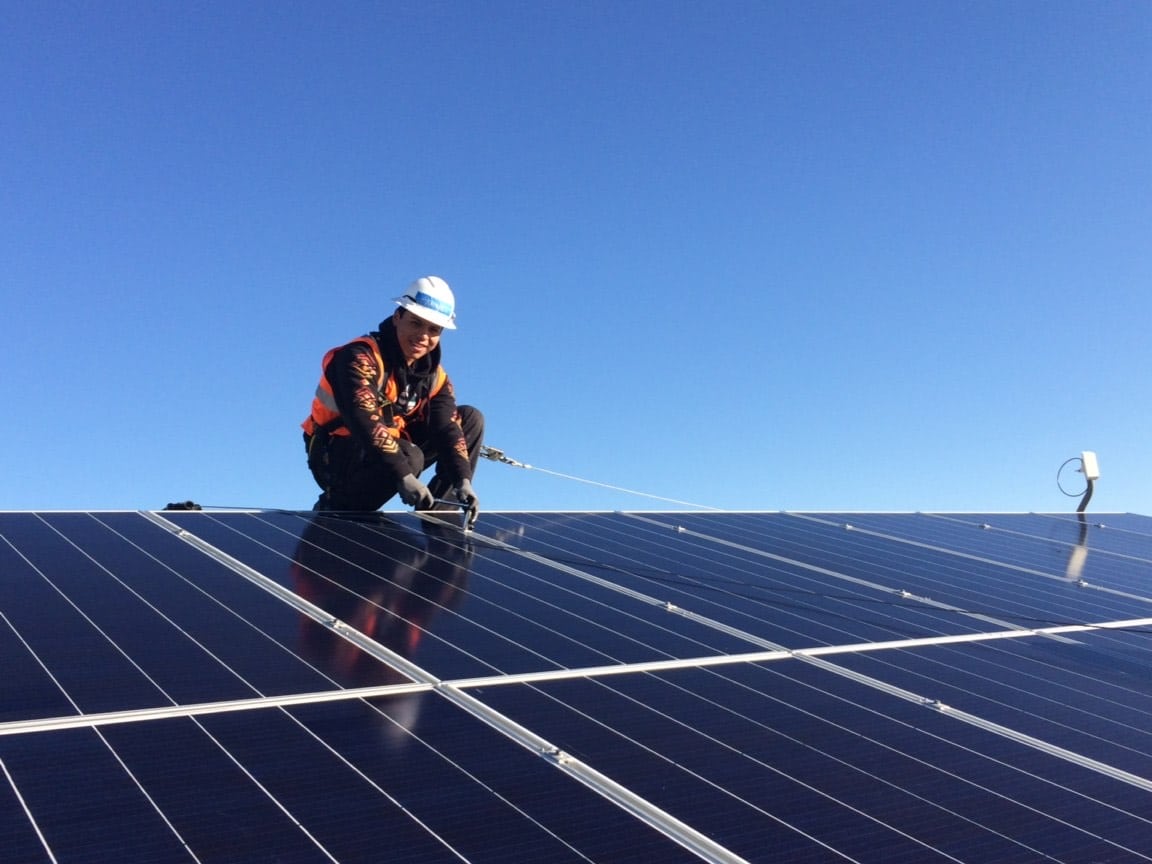 A trainee wearing an orange vest smiles while securing a solar panel on top of a roof. The sky is blue behind him.