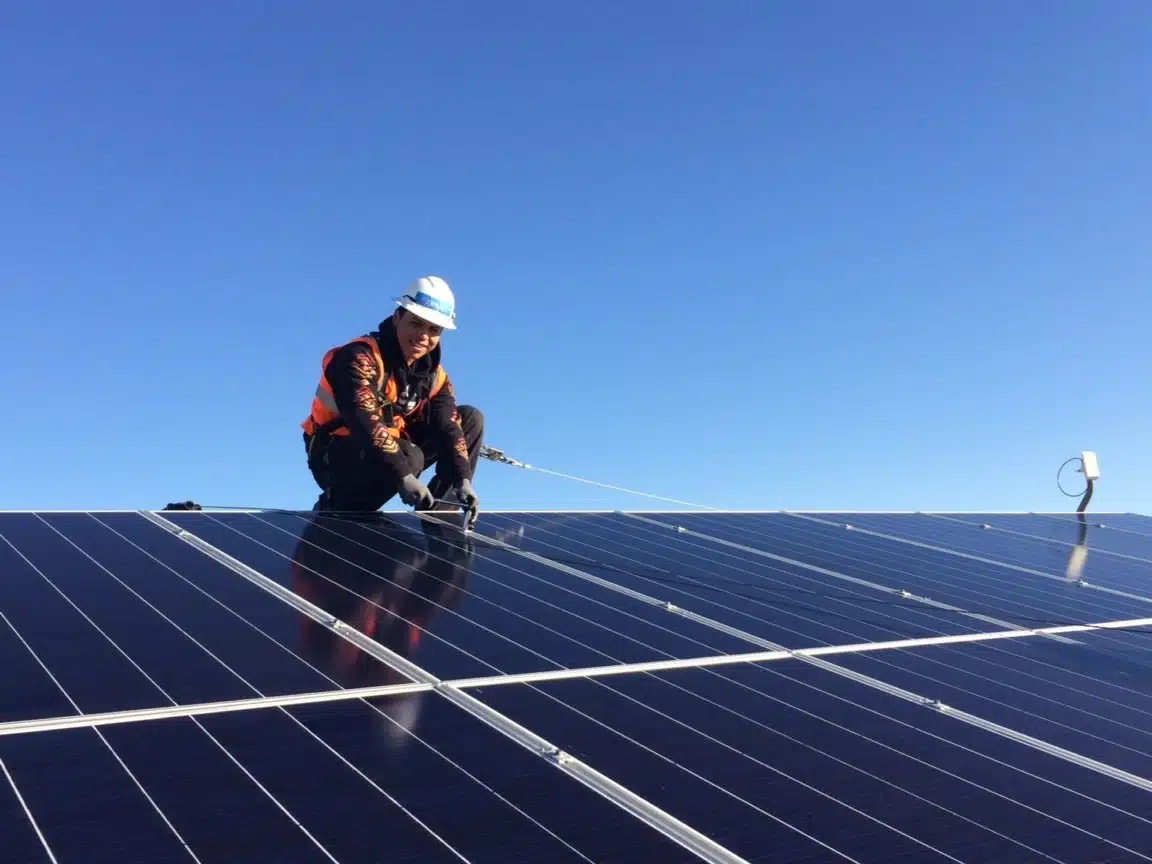 A trainee wearing an orange vest smiles while securing a solar panel on top of a roof. The sky is blue behind him.