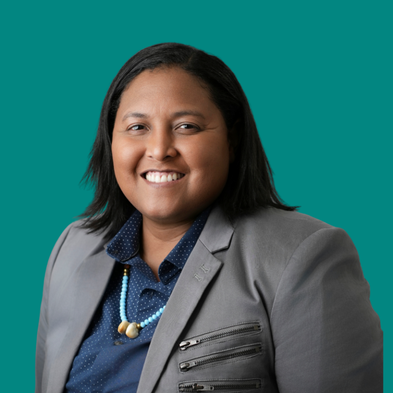 Professional headshot of Talia Martin smiling against turquoise background, wearing gray blazer and blue polka dot shirt with beaded necklace