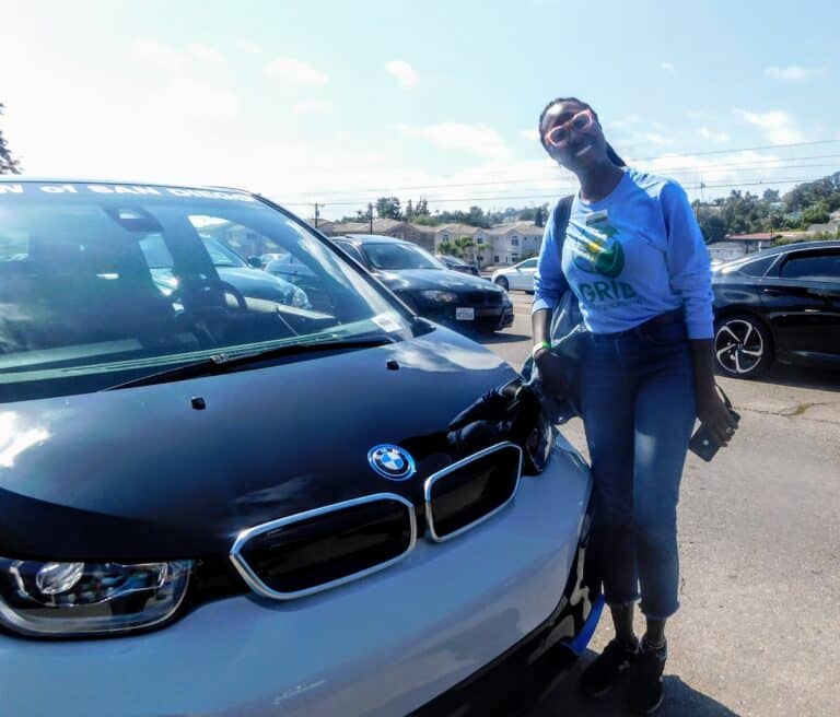 Person in blue "Grid" sweatshirt posing with white BMW i3 electric vehicle at outdoor event