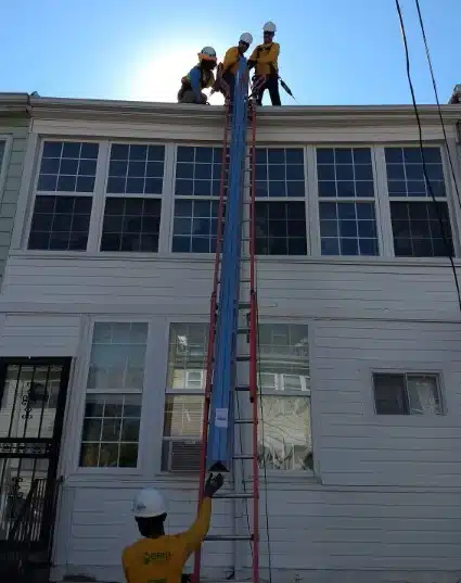 Three workers on a roof using a rope and pulley system to lift a blue rail up the side of a white building