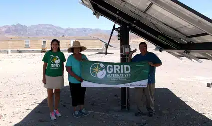 Four people holding a GRID Alternatives banner in front of solar panels in a desert landscape