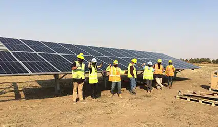 Group of workers in yellow safety vests standing beneath large solar panels in a desert setting