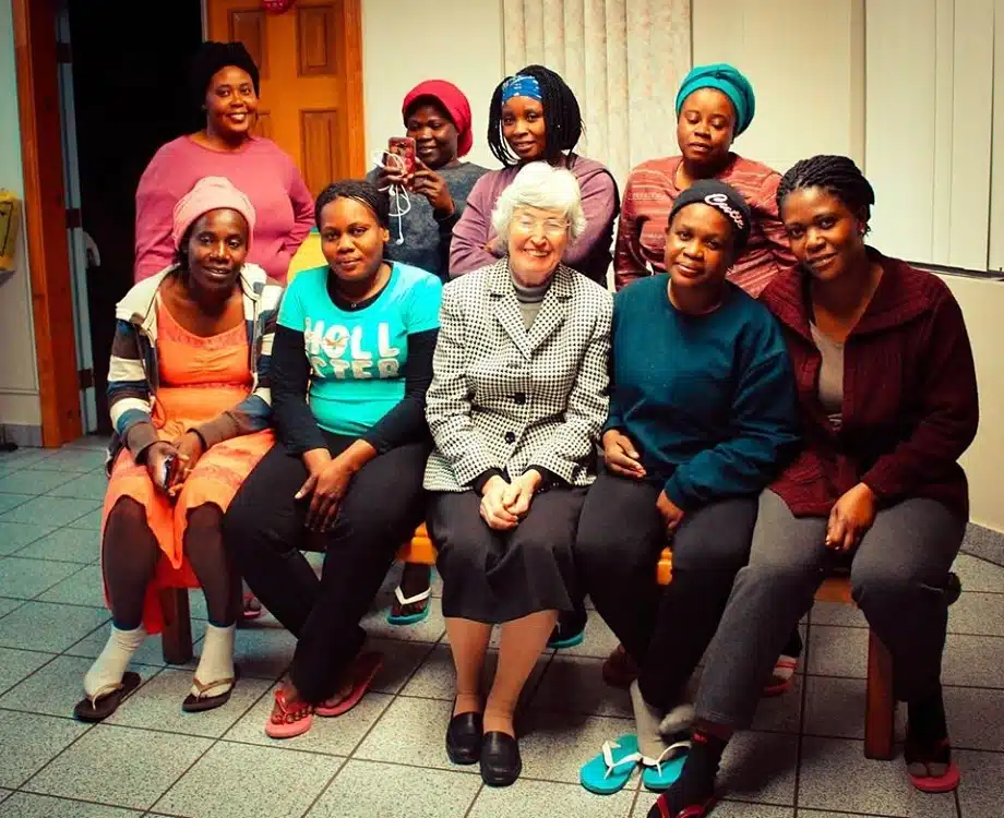 Group of nine women posing together indoors, smiling at camera in casual clothing and colorful headwraps