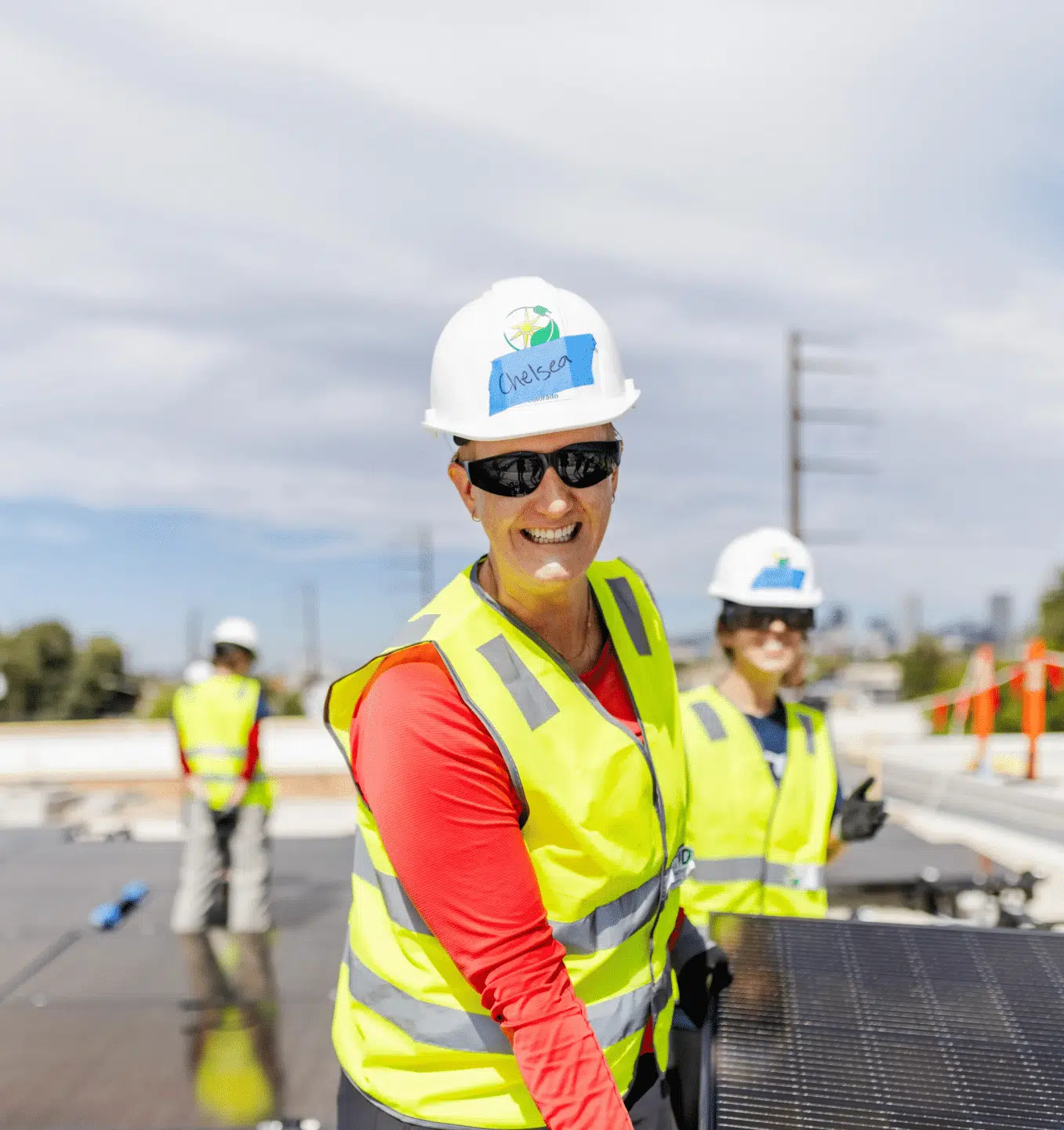 A GRID installer in a hard hat and safety vest smiles over her shoulder while working on a rooftop solar array.