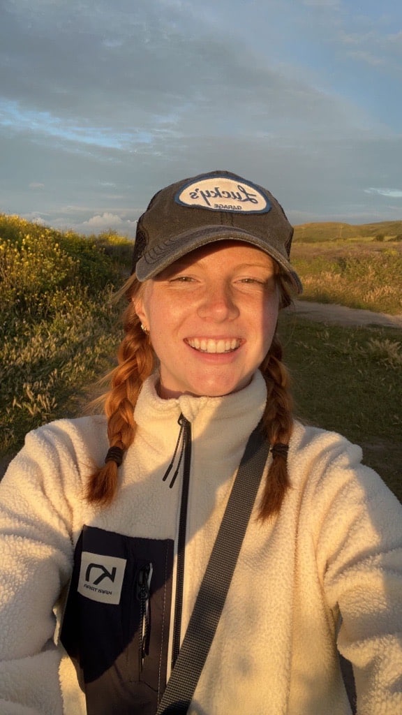 Smiling selfie of Emma at dusk in a field. They wear a baseball cap with a Lucky's logo, their auburn shoulder-length hair is tied into 2 braids.