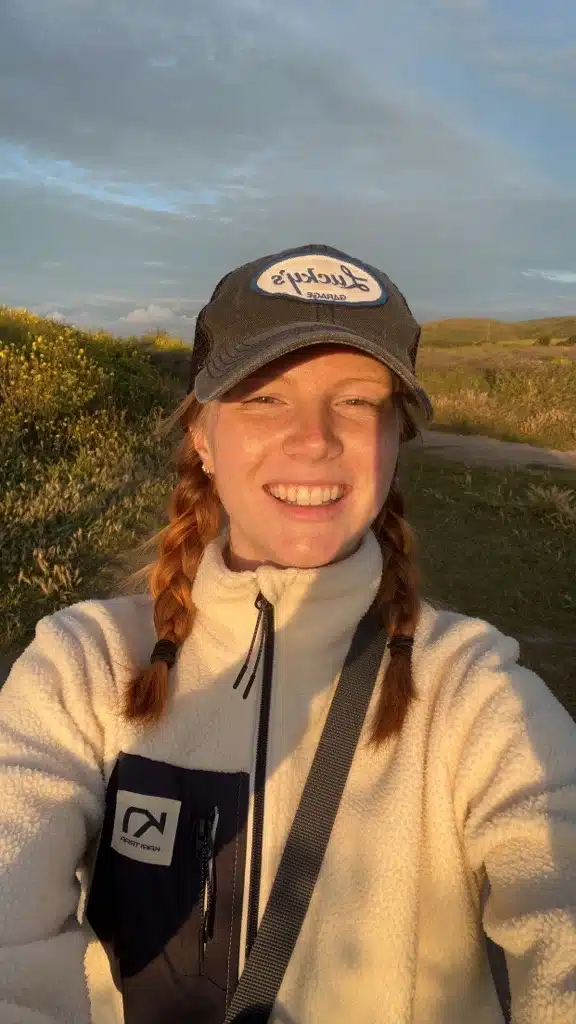 Smiling selfie of Emma at dusk in a field. They wear a baseball cap with a Lucky's logo, their auburn shoulder-length hair is tied into 2 braids.