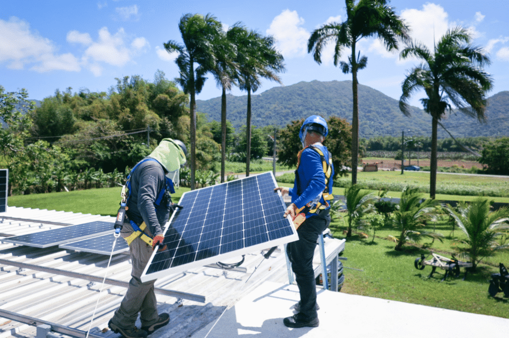 Two installers on a tropical rooftop carrying a solar panel into place, palm trees and mountains behind them.