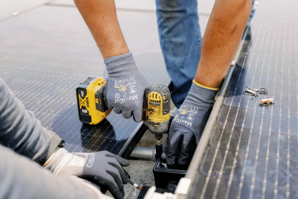 Close-up of two installers' gloved hands driving a screw into a rooftop solar panel mounting rail with a cordless drill.
