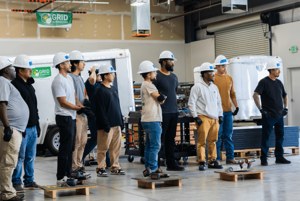 A line of trainees in hard hats and safety glasses standing at attention in a training warehouse beside a GRID Alternatives banner and equipment trailers.