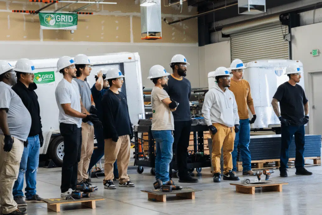 A line of trainees in hard hats and safety glasses standing at attention in a training warehouse beside a GRID Alternatives banner and equipment trailers.