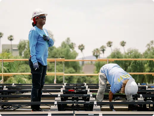 Two construction workers installing solar panels on a rooftop with palm trees in background