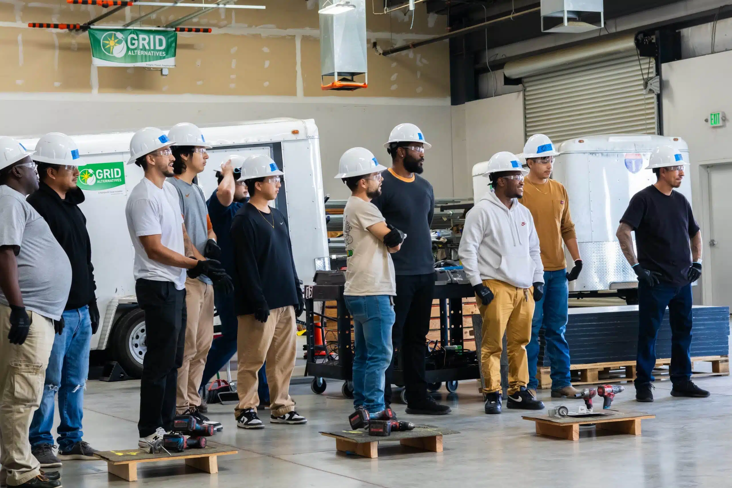 A cohort of GRID trainees in hard hats and safety glasses stands in a training warehouse, cordless drills and practice roof decks on the floor in front of them.