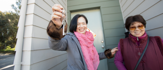 Two women holding keys outside a house with gray siding and green door