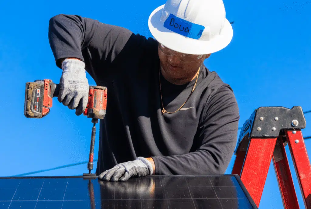 Worker in hard hat installing solar panel with tools against blue sky