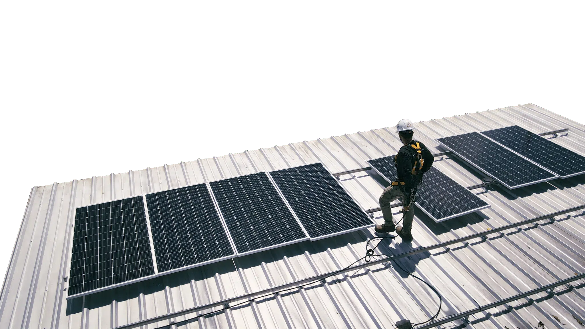 An installer in a hard hat and fall-arrest harness walks a metal roof between rows of newly mounted solar panels.