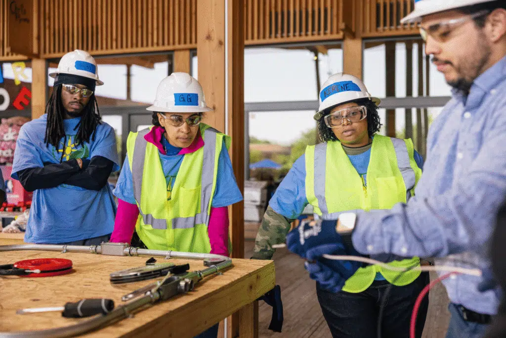 Construction trainees in hard hats and safety vests listen to instructor during hands-on building training session