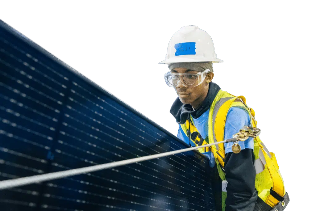 A trainee in a hard hat, safety goggles and a fall-arrest harness guides a solar panel into position.