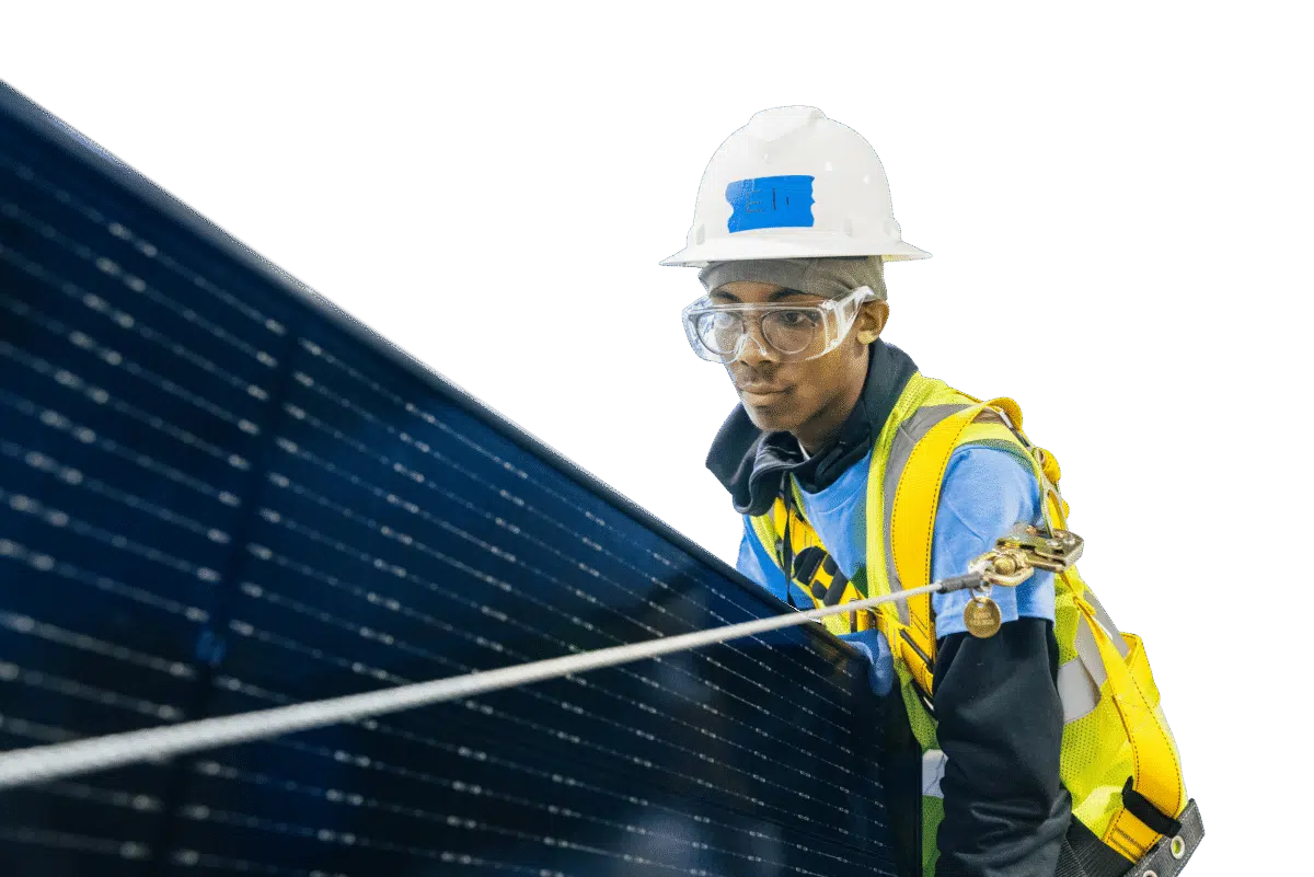 A trainee in a hard hat, safety goggles and a fall-arrest harness guides a solar panel into position.