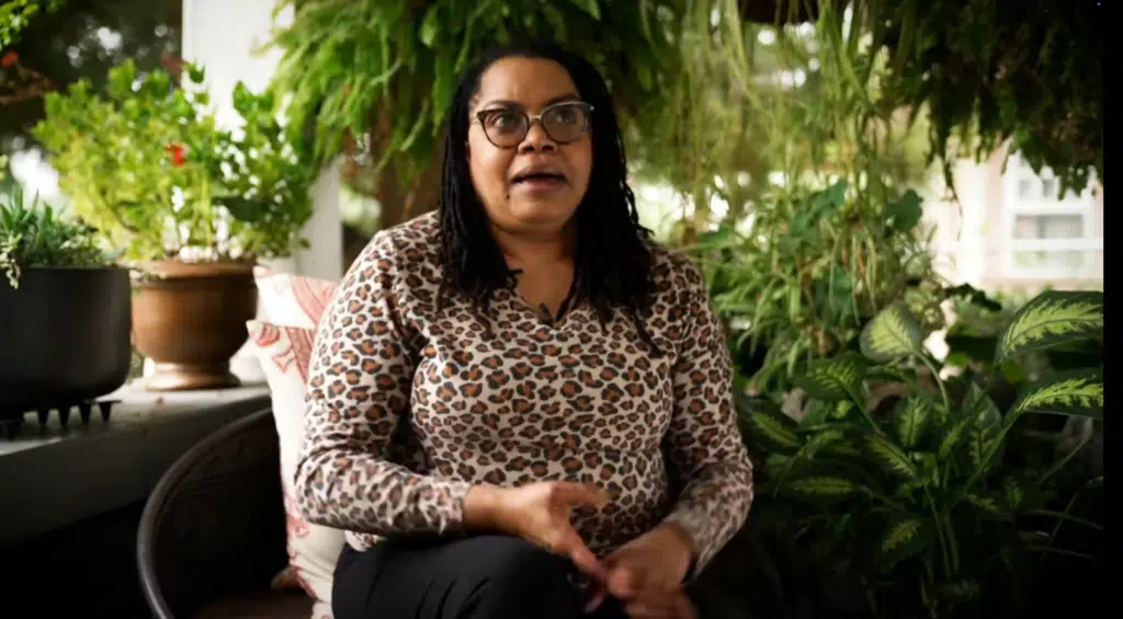 A woman seated on a plant-filled porch speaks to camera for a video testimonial.