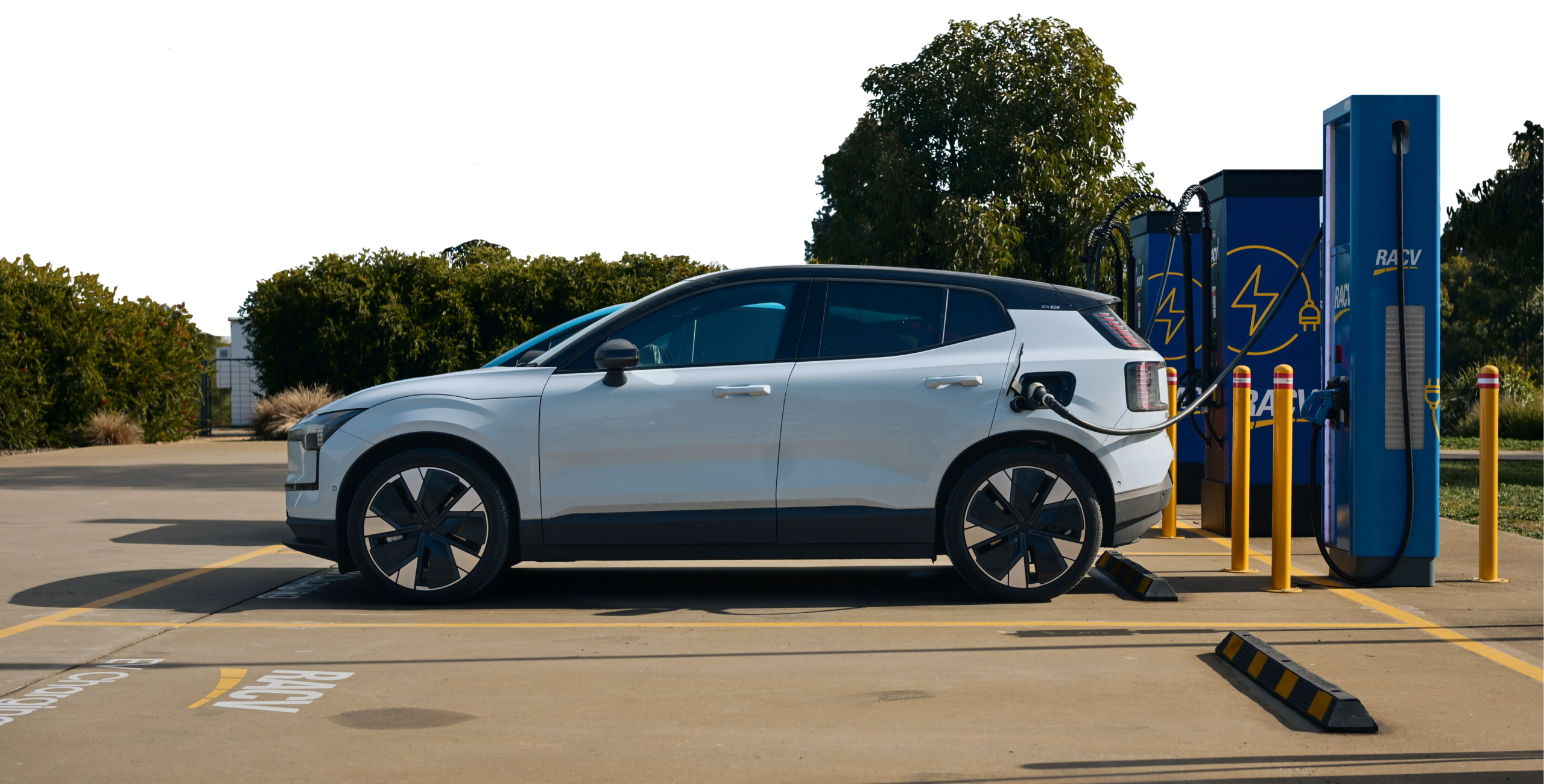 A white electric vehicle charges at a blue public RACV charging station surrounded by trees.