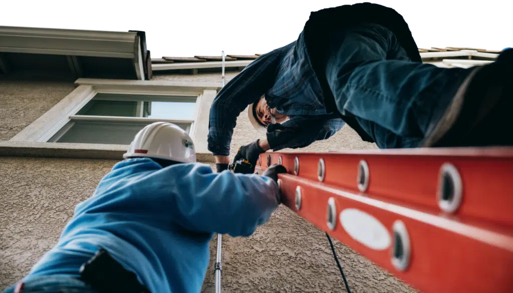 Two installers pass a ladder up the side of a building from a low, dramatic angle.