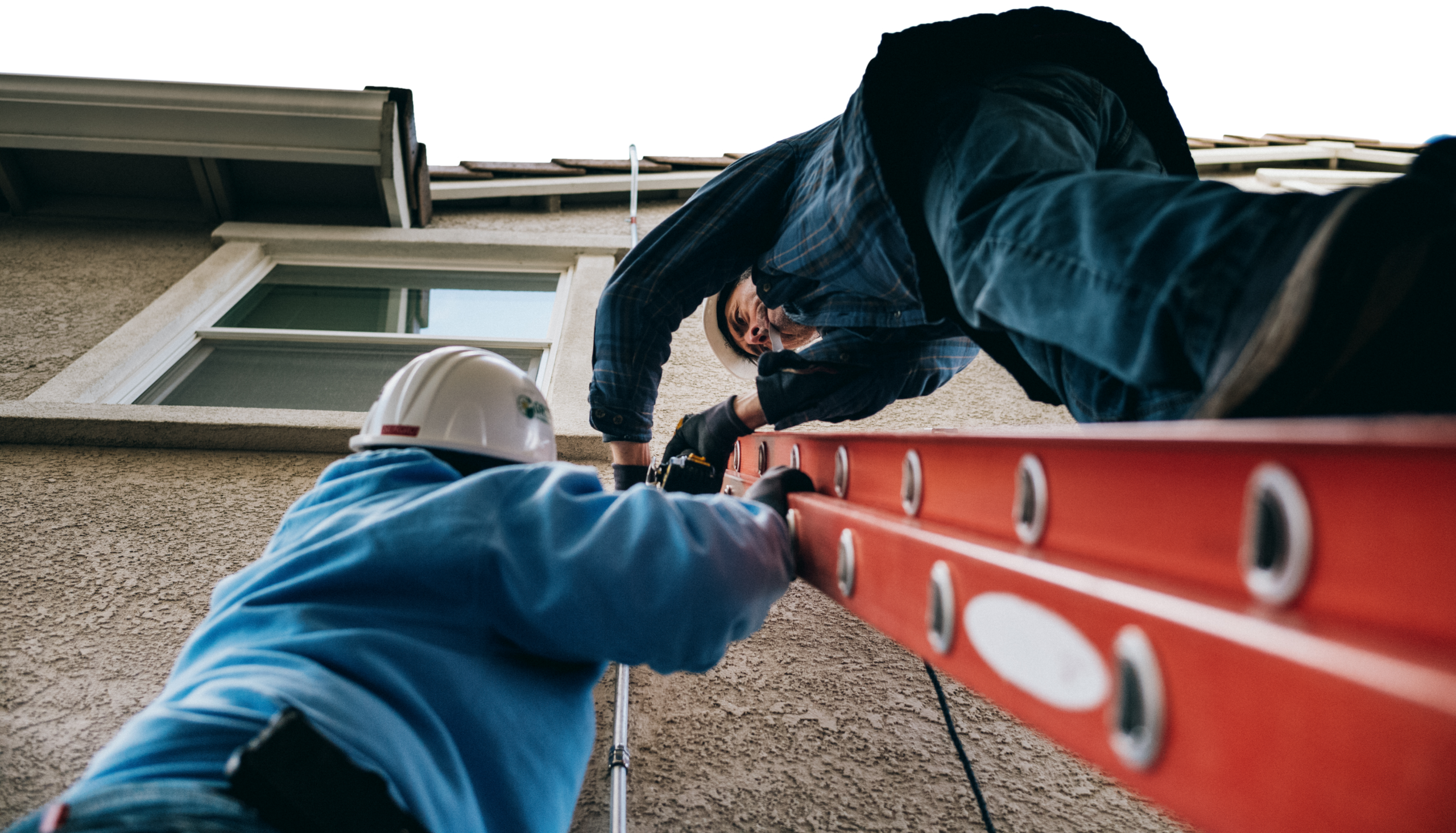 Two installers pass a ladder up the side of a building from a low, dramatic angle.