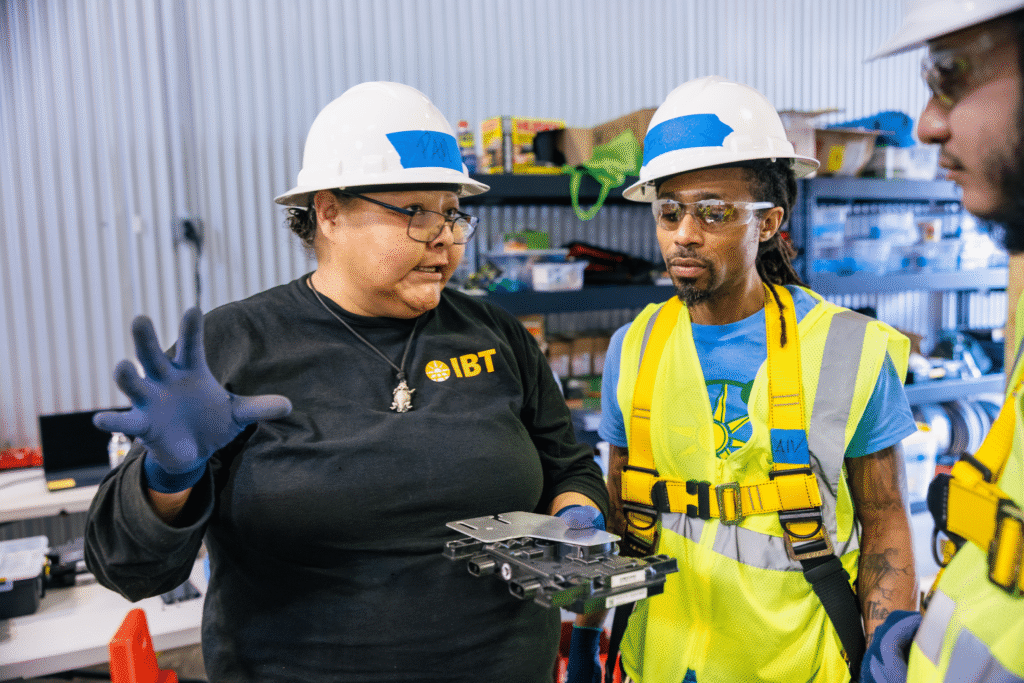 An IBT trainee wearing safety glasses gestures while explaining a wiring component to two classmates.