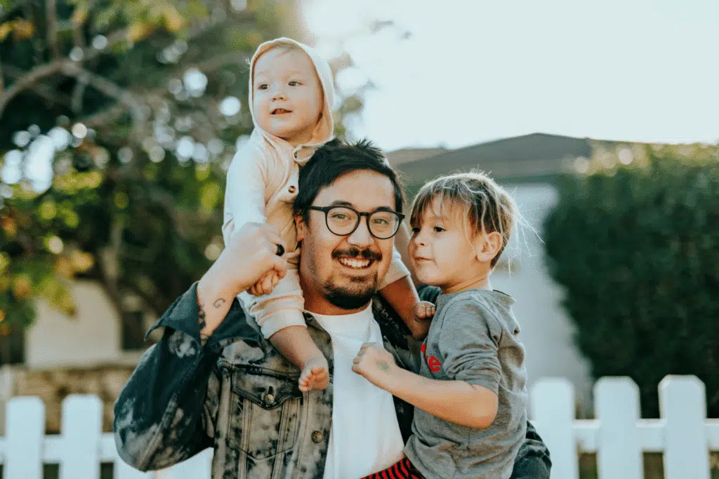 A father carries two children on his shoulders in a backyard.
