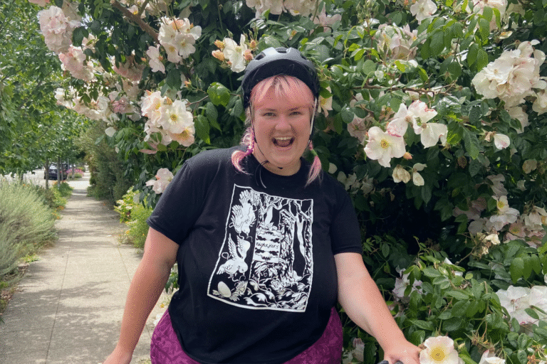 A woman with pink hair rides a bicycle past a bed of orange flowers.
