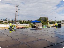 Installers in hard hats and safety vests working on a large flat rooftop solar array, with a canopy tent and city skyline in the background.