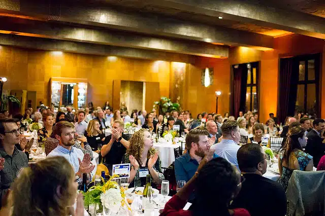 Large formal banquet hall with hundreds of guests seated at white-clothed tables with yellow floral centerpieces