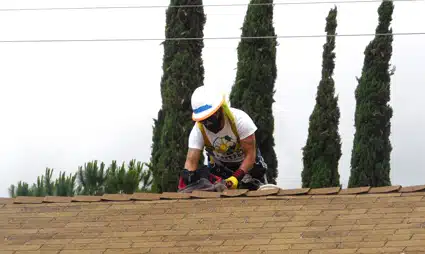 Worker in safety gear installing roofing material on a residential roof with cypress trees in background