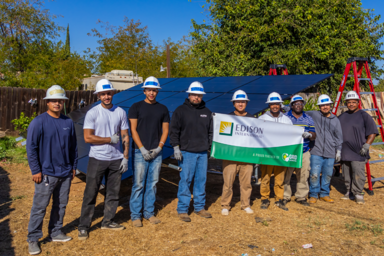 Nine installers and volunteers in hard hats standing behind a ground-mounted solar array, two of them holding an Edison International banner reading A proud partner of GRID Alternatives.