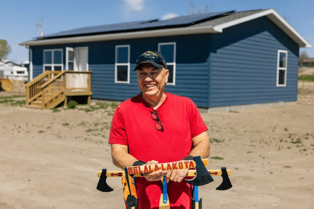 A smiling man in a red shirt holding two ceremonial tomahawks inscribed Oglala-Lakota, standing in front of a house with a newly installed rooftop solar array.