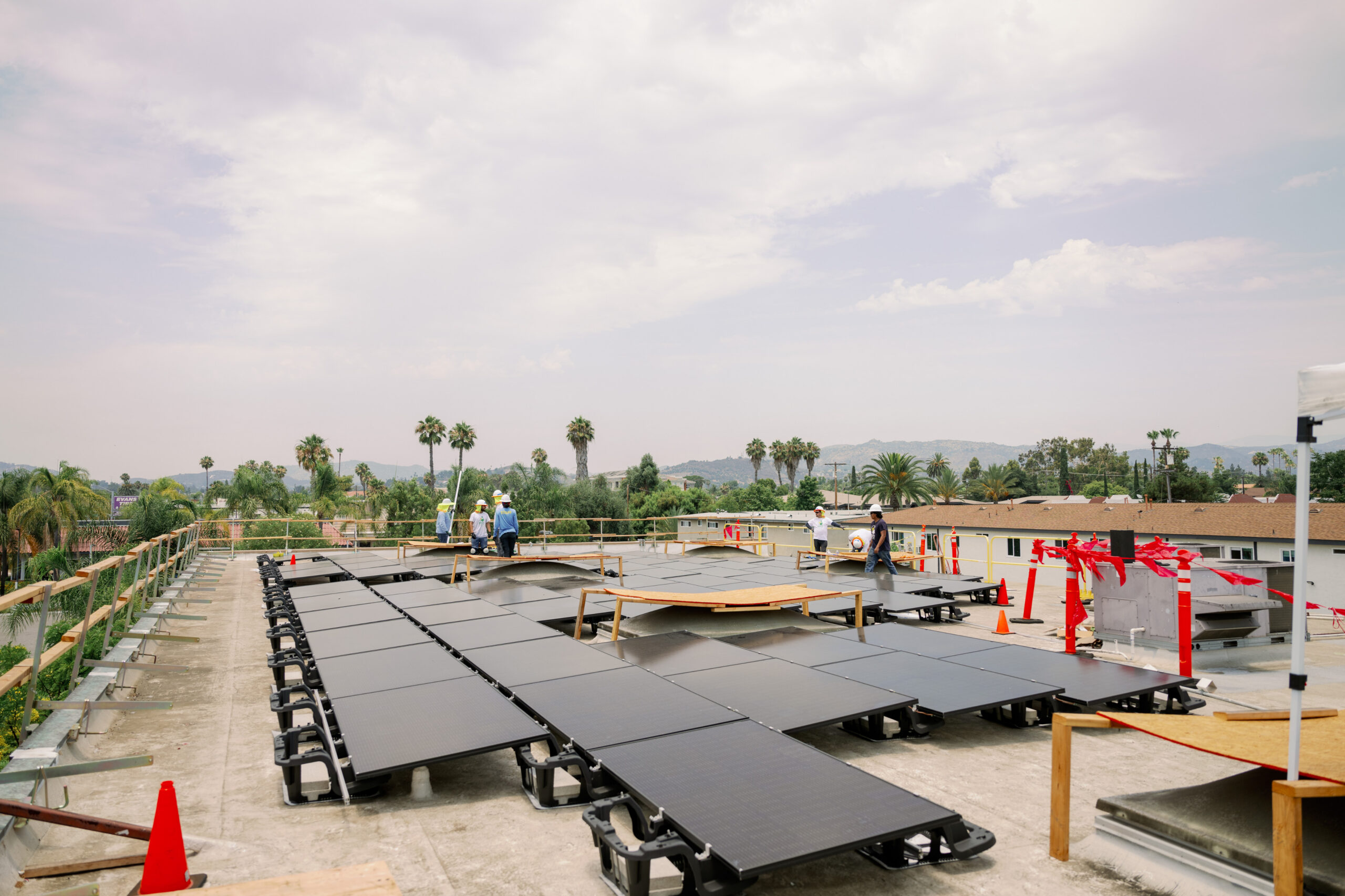 An overcast rooftop mid-installation, rows of ballasted solar panels laid out with a crew working nearby, palm trees and hills in the distance.