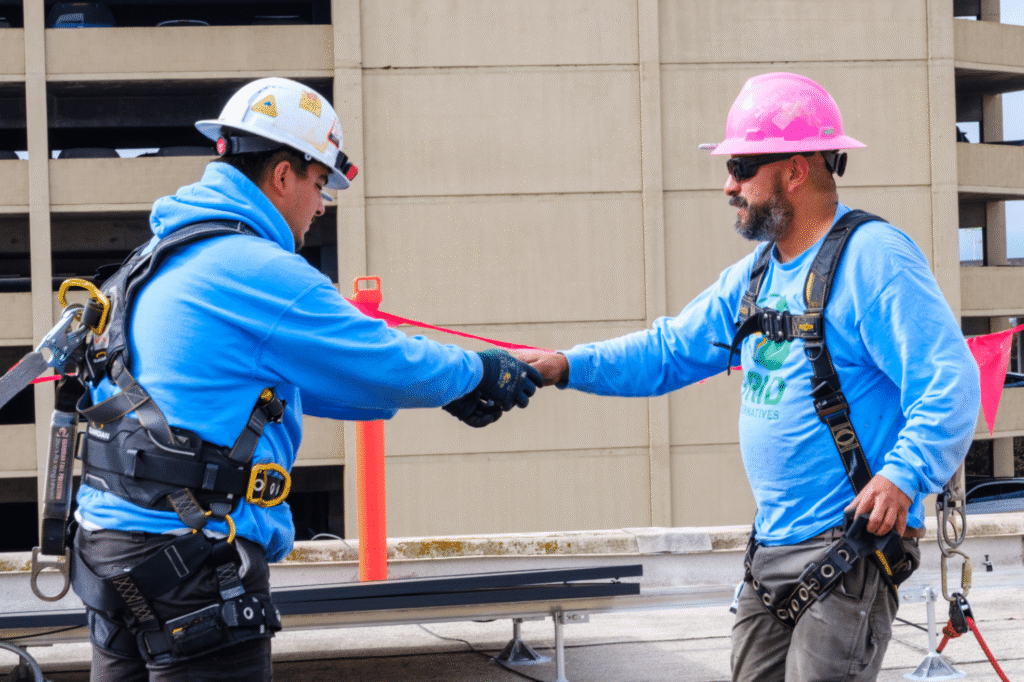 Two installers in hard hats passing a red strap between them on a rooftop, a parking structure behind.